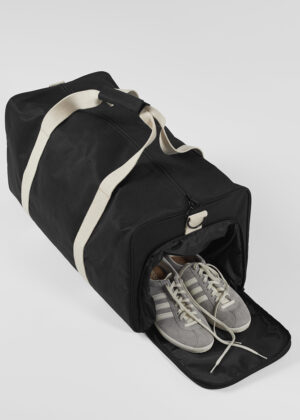 Black duffel bag with beige straps and a zipped top, shoes stored in the front compartment with the door open to reveal gray sneakers inside.