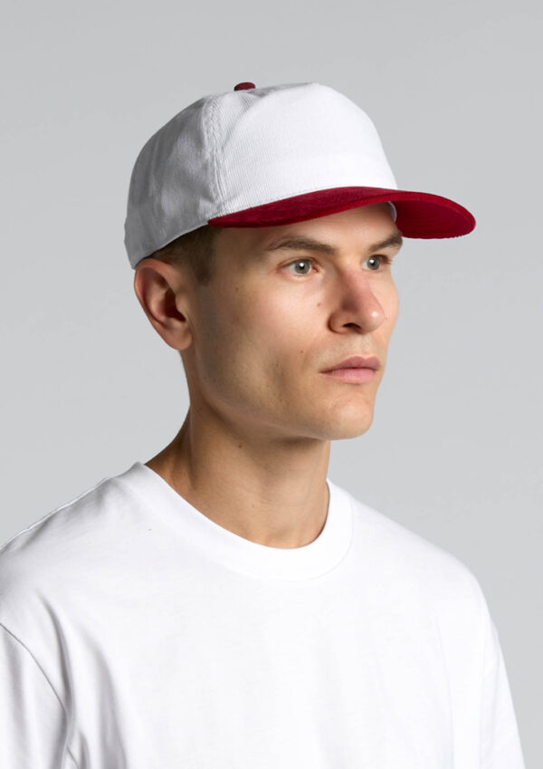 Portrait of a man wearing a white baseball cap with a red brim, and a white crew-neck shirt, facing slightly to the right against a gray background.