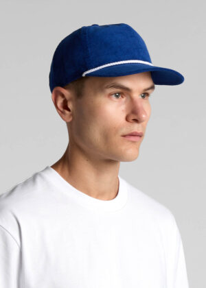 Man wearing a blue baseball cap with a white rope detail and a white T-shirt, facing right in a neutral studio background.