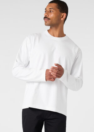 Man in a white long-sleeve shirt standing, looking to his left with hands loosely clasped in front.