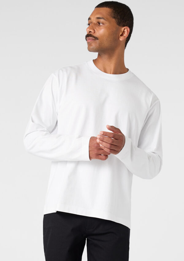Man in a white long-sleeve shirt standing, looking to his left with hands loosely clasped in front.
