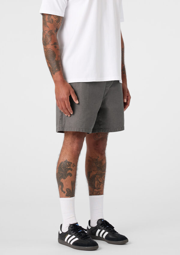 Full-body shot of a person in a white T-shirt, gray shorts, white socks and black sneakers with white stripes; tattoos visible on arms and legs.