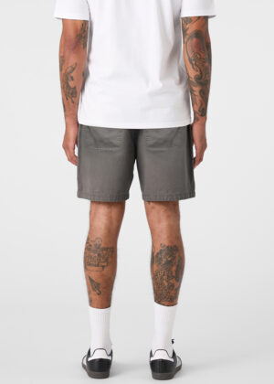 Back view of a person wearing a white T-shirt, gray shorts, white socks, and black sandals; tattoos cover both arms and legs visible from behind.