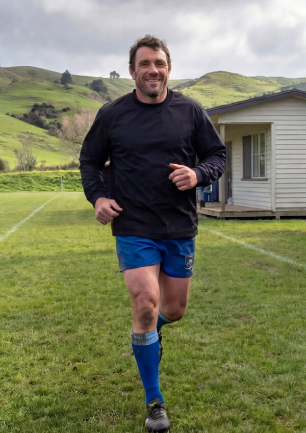 Smiling man running toward the camera on a grassy sports field, wearing a black long-sleeve shirt and blue shorts with blue socks in front of a small building and green hills.