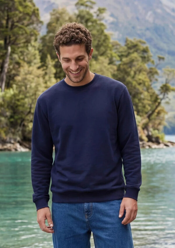 Smiling man in a navy sweatshirt and jeans standing by a turquoise lake with trees and mountains in the background.