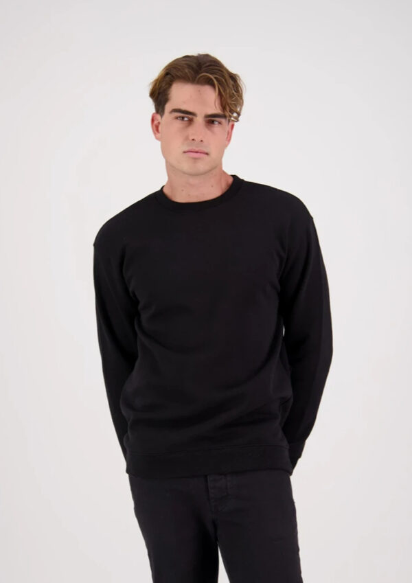 Young man in a black crewneck sweatshirt and black pants standing against a light studio background, looking straight at the camera.