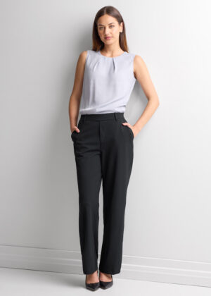 Woman standing against a light wall in a sleeveless gray blouse and black dress pants with hands in pockets.