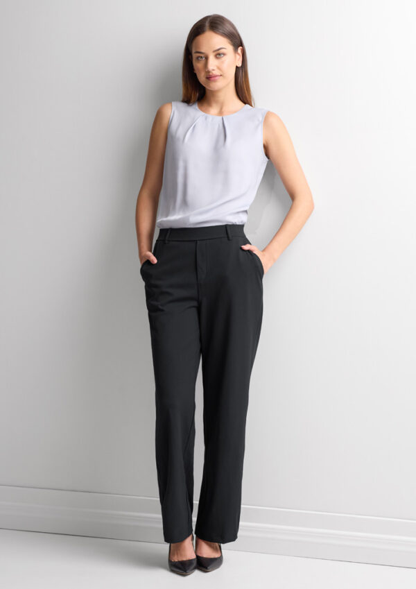 Woman standing against a light wall in a sleeveless gray blouse and black dress pants with hands in pockets.