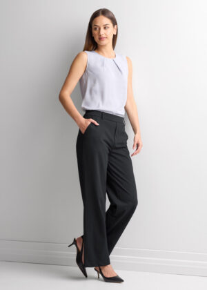 Woman stands in profile against a light wall, wearing a sleeveless light gray blouse, black wide-leg pants, and black high heels.