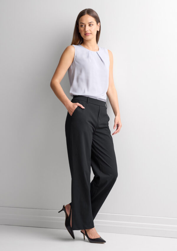 Woman stands in profile against a light wall, wearing a sleeveless light gray blouse, black wide-leg pants, and black high heels.