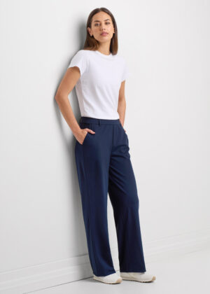 Woman wearing a white fitted T-shirt and navy wide-leg trousers, standing against a white wall with hands in pockets.
