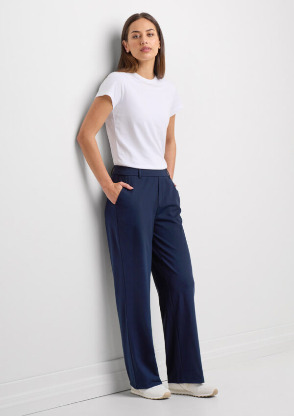 Woman wearing a white fitted T-shirt and navy wide-leg trousers, standing against a white wall with hands in pockets.