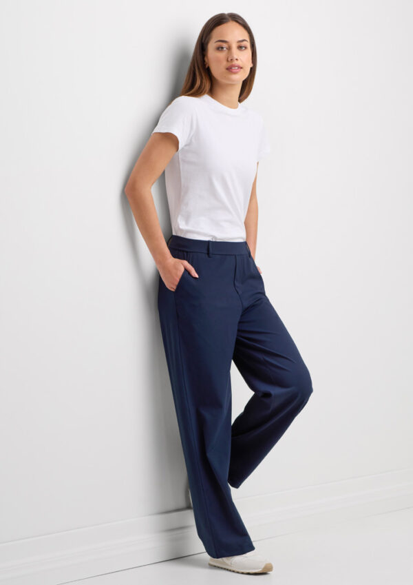 Woman in a white t-shirt and navy wide-leg pants leaning against a white wall with hands in pockets.