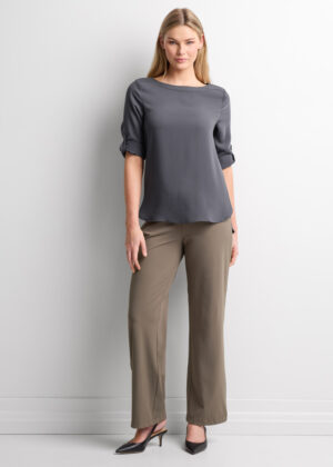 Woman standing against a light wall, wearing a gray blouse with rolled sleeves and taupe dress pants with black heels.