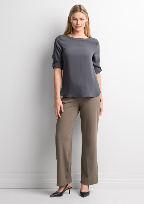 Woman standing against a light wall, wearing a gray blouse with rolled sleeves and taupe dress pants with black heels.