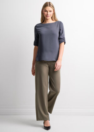 Woman wearing a blue-gray satin blouse with rolled sleeves and wide-leg taupe pants, standing against a white backdrop.
