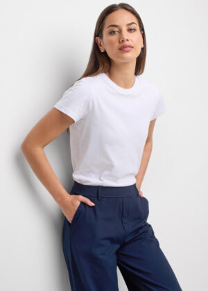 Woman in a white t-shirt and navy pants leaning against a wall with hands in pockets.