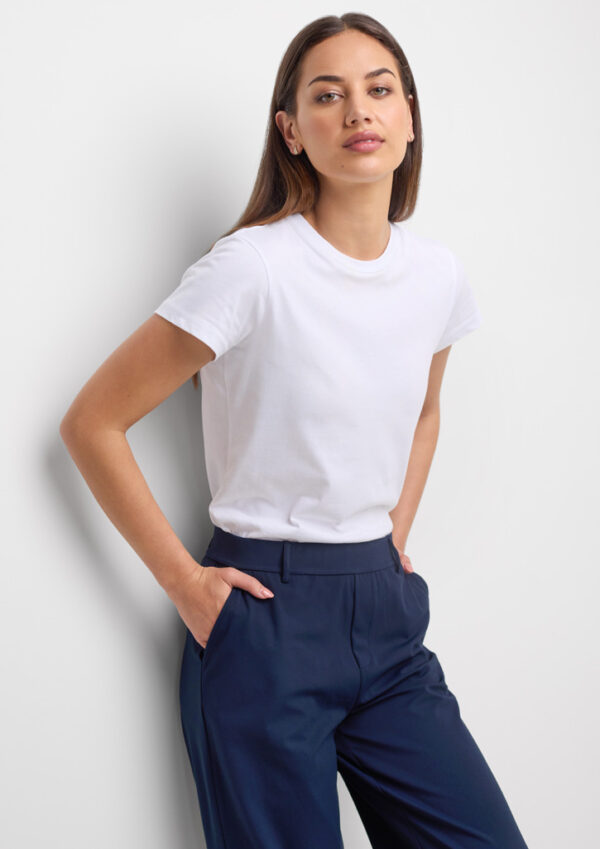 Woman in a white t-shirt and navy pants leaning against a wall with hands in pockets.