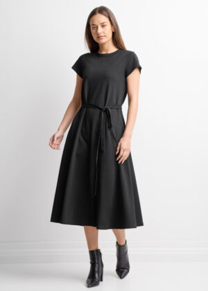 Woman in a black midi dress with a waist tie, paired with black ankle boots, standing against a white backdrop to the camera.