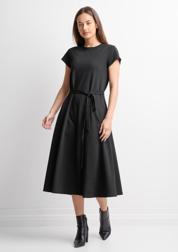Woman in a black midi dress with a waist tie, paired with black ankle boots, standing against a white backdrop to the camera.