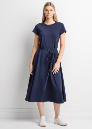 Woman standing in a navy midi dress with a self-tabric belt tied at the waist, wearing white sneakers against a white wall.