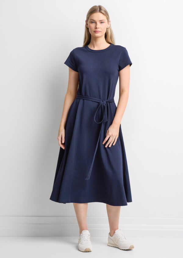 Woman standing in a navy midi dress with a self-tabric belt tied at the waist, wearing white sneakers against a white wall.