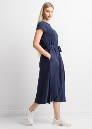 Model wearing a navy midi dress with a belt and pockets, standing sideways against a white background, white sneakers visible.