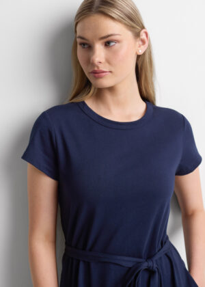 Young woman with blonde hair wearing a navy short-sleeve dress with a tied waist belt, standing against a light wall.