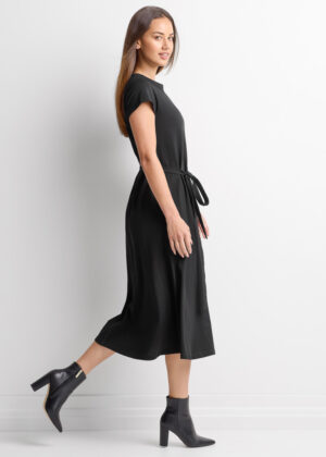 Woman in a black midi dress with a tie belt, walking in black ankle boots against a white background.