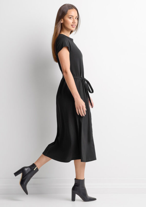 Woman in a black midi dress with a tie belt, walking in black ankle boots against a white background.