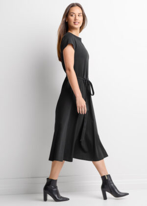 Side view of a woman walking, wearing a black midi dress with a matching belt and black ankle boots against a white studio background, minimal jewelry.