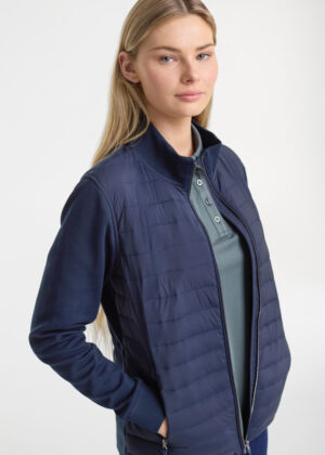 Woman wearing a navy quilted jacket with hands in pockets, facing slightly to the side, in a studio setting.