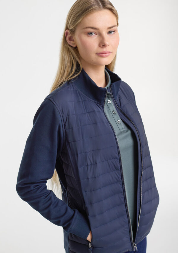 Woman wearing a navy quilted jacket with hands in pockets, facing slightly to the side, in a studio setting.
