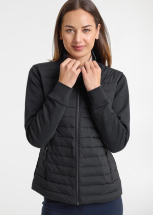 Woman wearing a black quilted zip-up vest, standing against a light background, hands near the collar as she adjusts it.