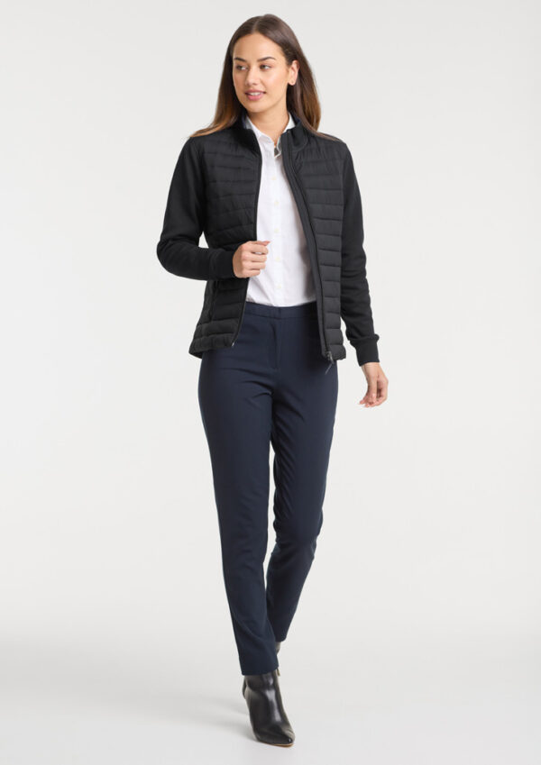 Full-body shot of a woman wearing a black quilted jacket over a white shirt, navy trousers, and black ankle boots.