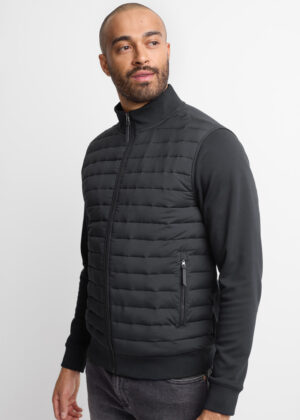 Man wearing a black quilted zip-up vest over a black long-sleeve shirt, standing in a studio pose with hands at sides.
