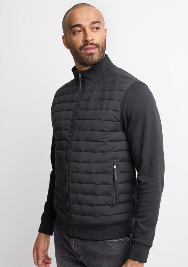 Man wearing a black quilted zip-up vest over a black long-sleeve shirt, standing in a studio pose with hands at sides.