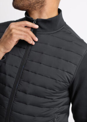 Man wearing a black quilted vest, zipping up the front zipper at the chest level.