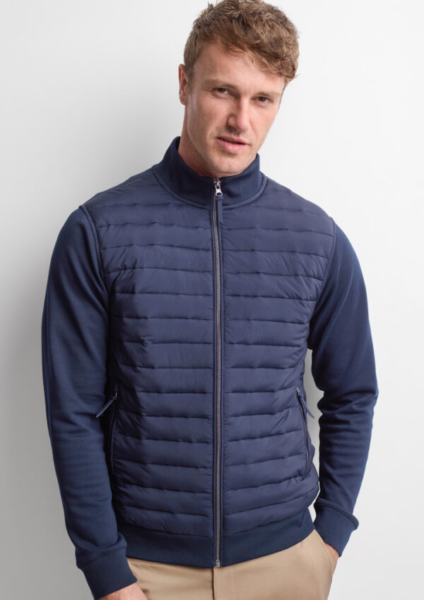 Man posing in a navy quilted zip-up vest over a navy long-sleeve shirt, standing against a white wall, beige pants visible at the bottom.]