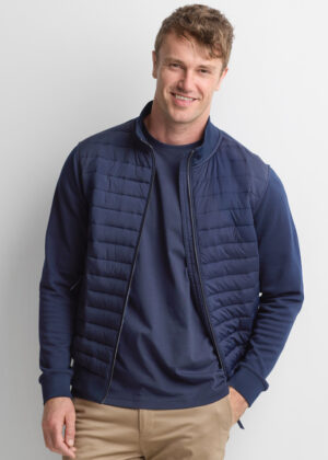Man smiling in a navy quilted vest over a matching navy long-sleeve shirt and beige khaki pants.