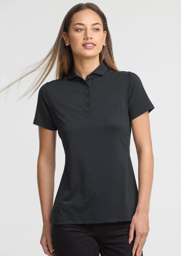 Woman wearing a black polo shirt with short sleeves and a button placket, standing against a light background.”,