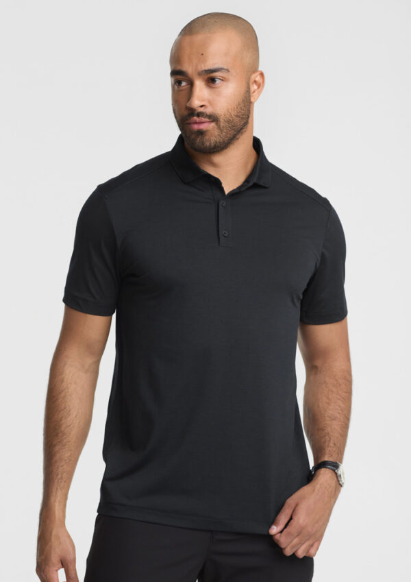 Male model wearing a plain black polo shirt, short sleeves, standing against a light gray background, facing slightly left.