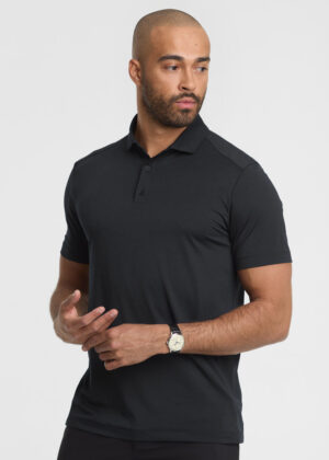 Man modeling a fitted black polo shirt with short sleeves, looking to the side, wearing a wristwatch against a light background.
