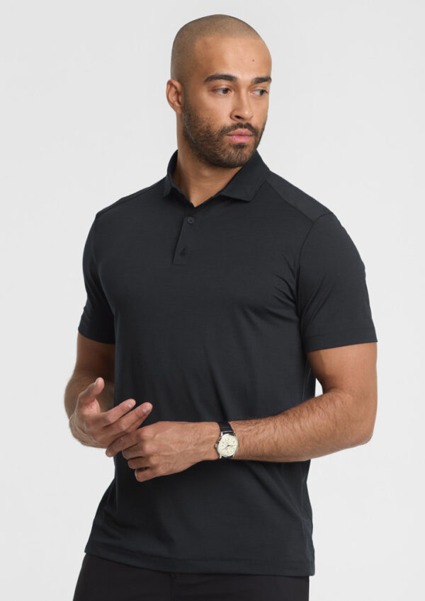 Man modeling a fitted black polo shirt with short sleeves, looking to the side, wearing a wristwatch against a light background.
