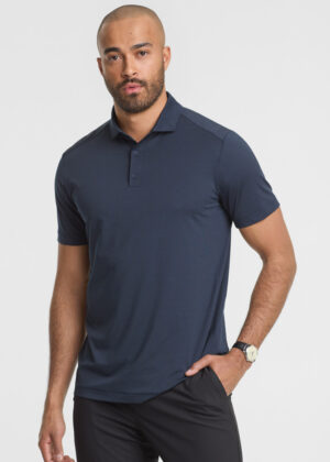 Man wearing a navy polo shirt and dark pants, standing with one hand in pocket against a light background.
