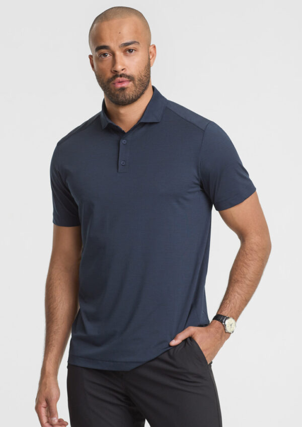 Man wearing a navy polo shirt and dark pants, standing with one hand in pocket against a light background.