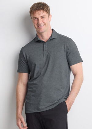 Smiling man wearing a gray polo shirt and dark pants, leaning against a white wall.