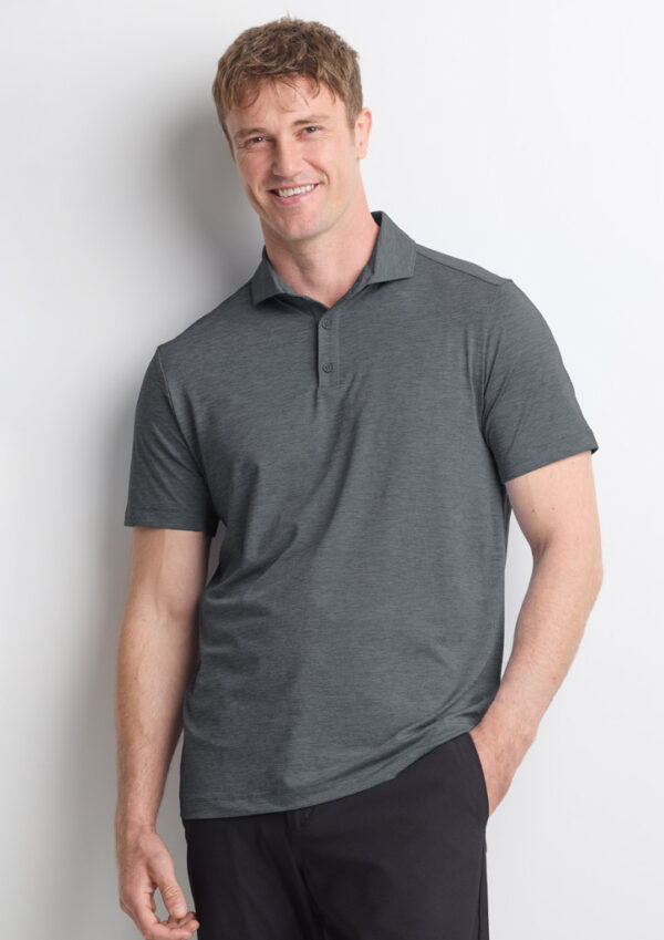 Smiling man wearing a gray polo shirt and dark pants, leaning against a white wall.