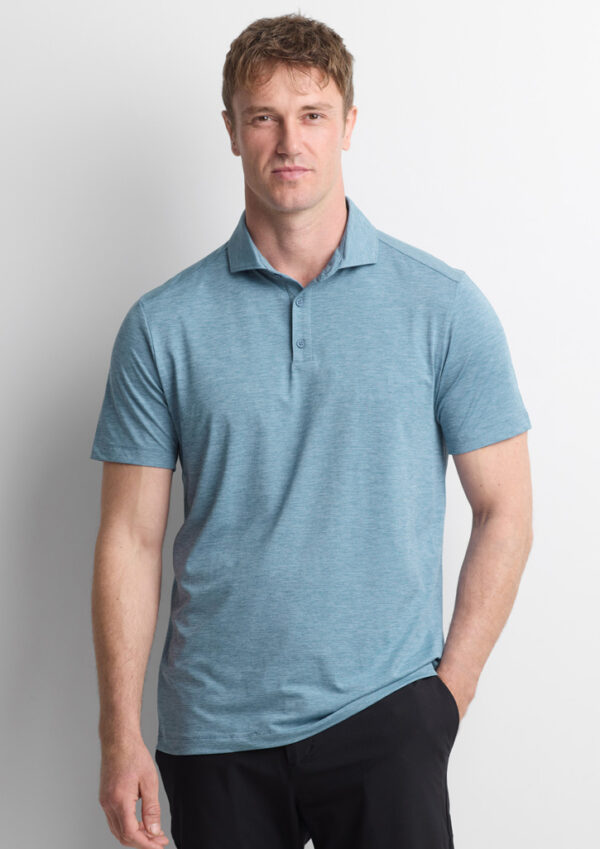 Portrait of a man wearing a light blue polo shirt standing against a white wall, with one hand in his pocket and a neutral expression.