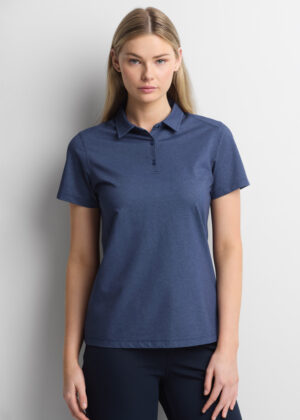 Woman wearing a blue polo shirt and navy pants, standing against a light gray wall.